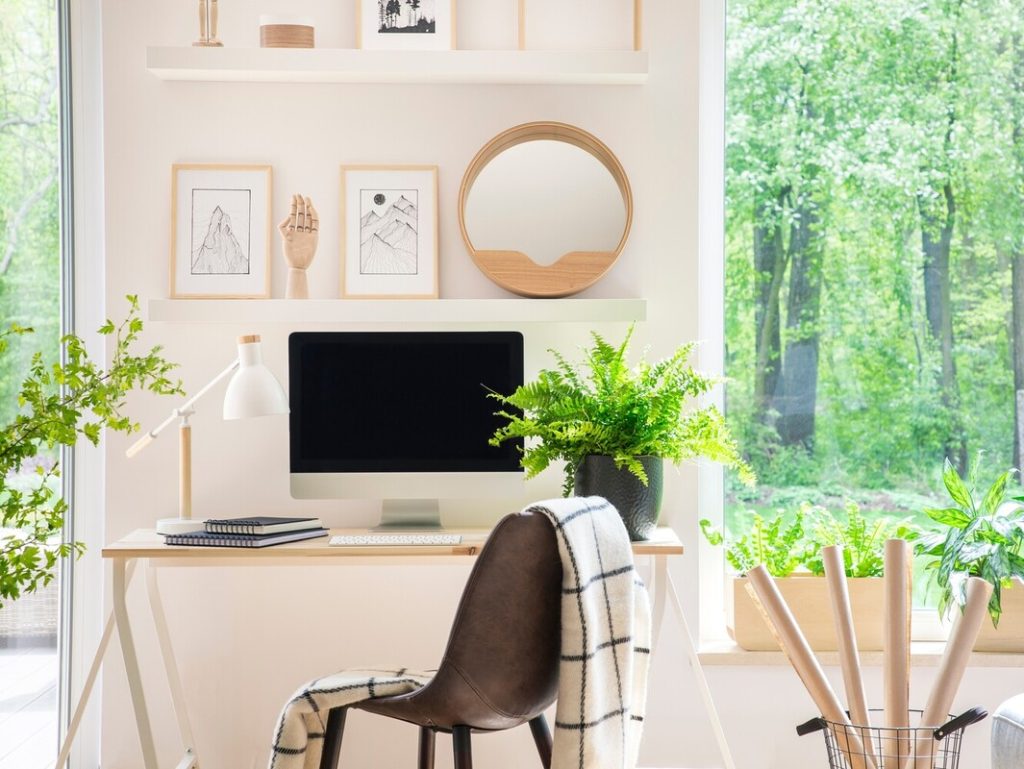 Shelves with illustrations above a wooden desk with computer by a window in a natural, white home office interior.