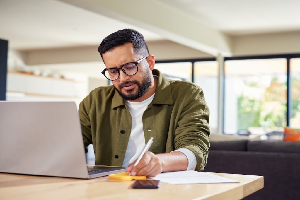 man writing on paper in front of laptop