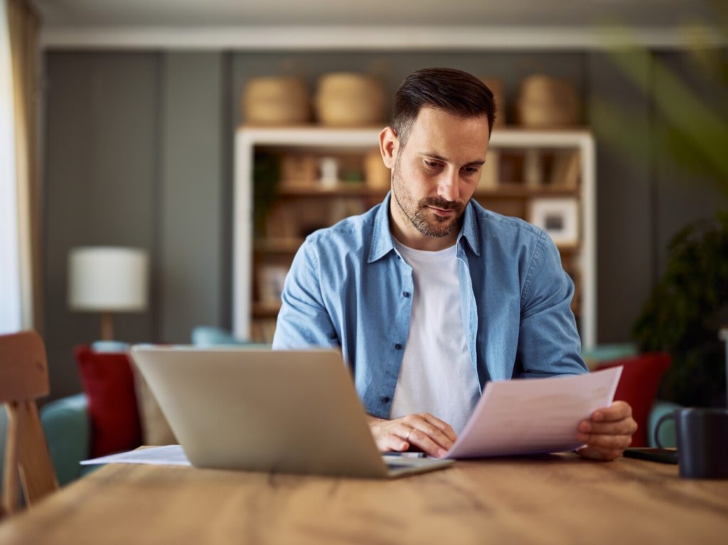 A focused adult freelance man reading a document and sitting in front of a laptop in a home office.
