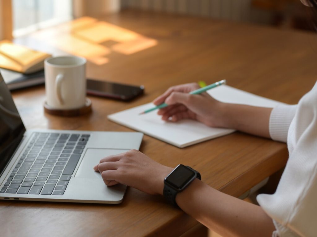 Cropped shot of young woman working on laptop while writing on notebook