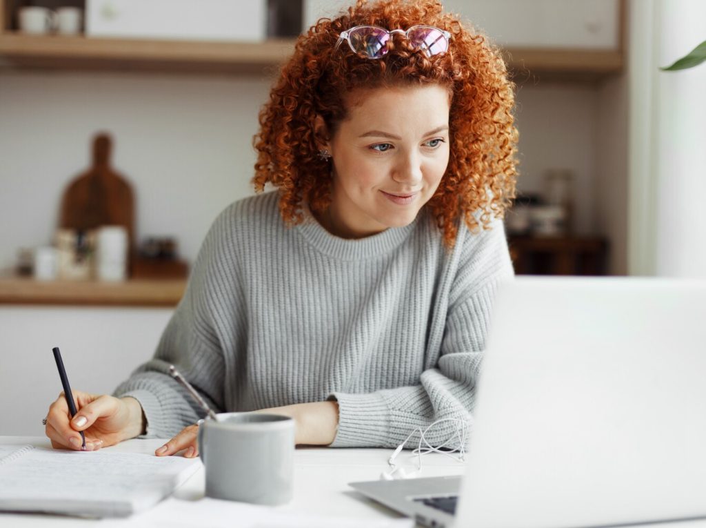 Portrait of happy female student watching webinar or tutorial online on laptop sitting at kitchen table.