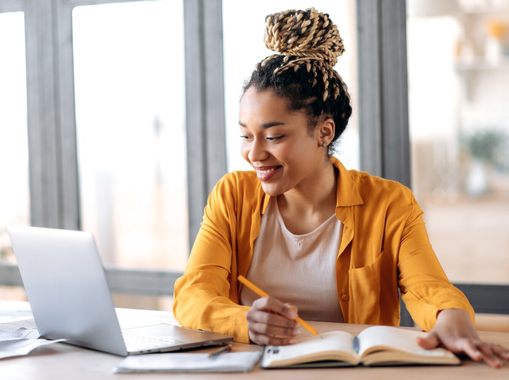 woman smiling and working from her laptop and taking notes in a book
